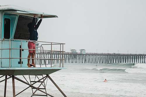 oceanside men's black pullover hoodie and red shorts reaching white metal frame beside sea during daytime ocean