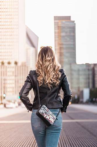 person woman wearing black leather jacket and blue denim bottoms standing on gray concrete surface near buildings during daytime people