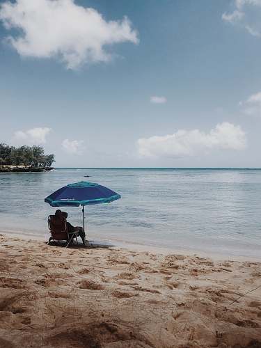 water person sitting on chair under blue umbrella on beach during daytime shoreline