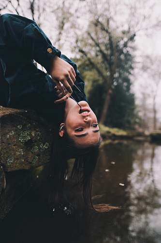 human selective focus photography of woman sitting on moss-covered stone with hair on river during daytime person