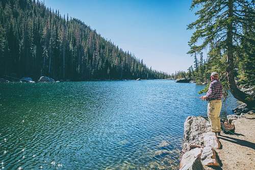 lake man standing on rock on front of lake surrounded with trees at daytime fishing