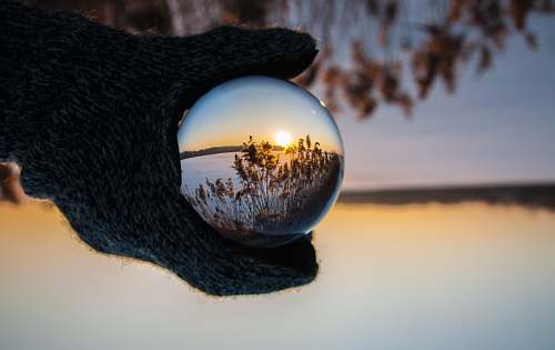 glass person wearing black gloves holding glass ball reflecting body of water with sunlight sunset