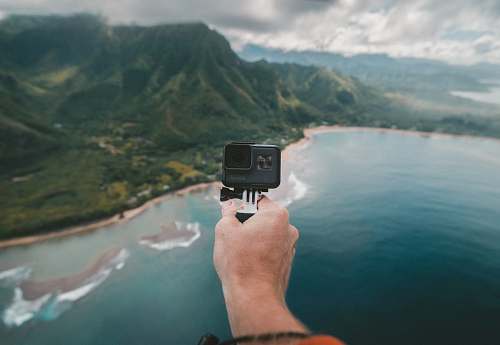 island person holding black action camera fronting green mountains ocean