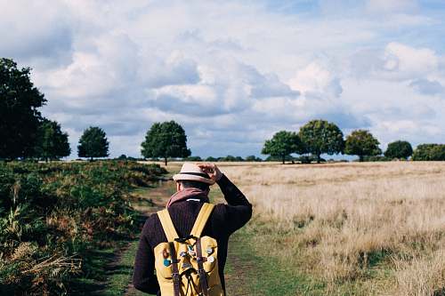 person man holding fedora cap while walking on green and beige grass field under white clouds people