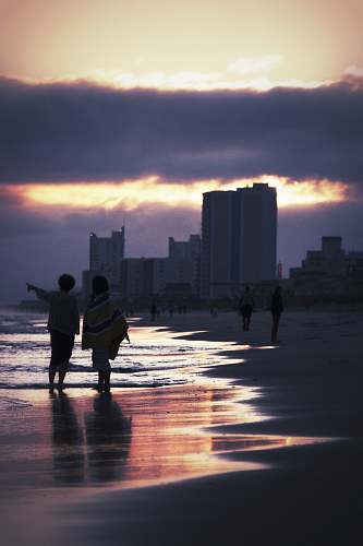 person two people walking on seashore near buildings during golden hour silhouette