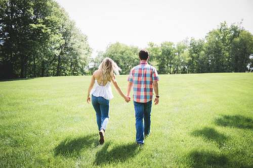 woman man and woman walking on green grass field surrounded with trees holding hands