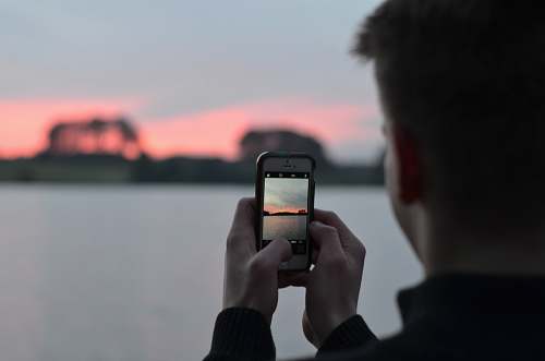 human selective focus photography of person using smartphone camera taking photo of blue body of water people