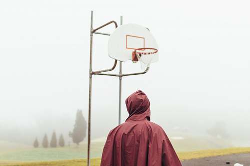 outdoors person wearing maroon raincoat standing under white and grey basketball hoop during foggy daytime outdoor