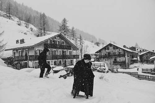 person grayscale photo of man covered with coat near houses snow
