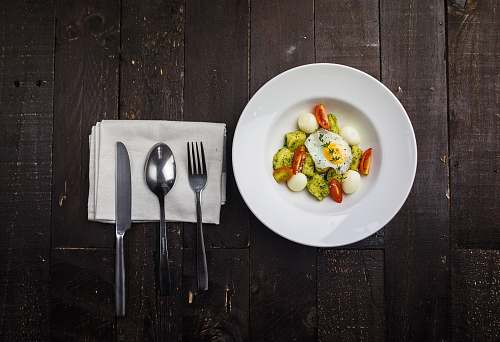 plate sunny side up egg, boiled quail eggs, sliced red tomatoes, and sliced green bell pepper inside round white ceramic plate in a fine dining set cutlery