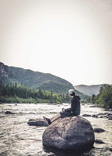 human man sitting on rock viewing river and mountain during daytime wilderness