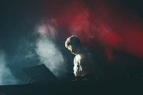 music A musician in a white shirt on stage with white and red lights breaking through the smoke behind his back man