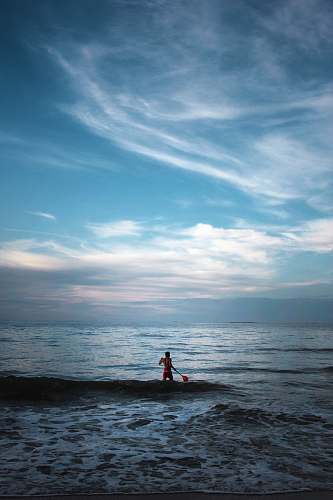 sky man standing on body of water holding paddle clouds