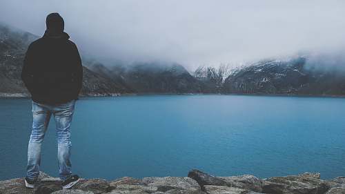 person man standing on cliff facing bod of water lake