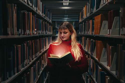 library woman in red turtleneck sweater holding book between library shelves indoors