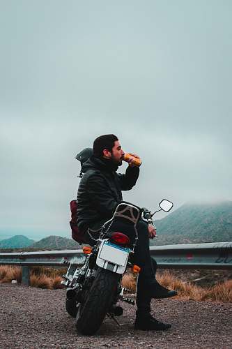 human man sitting on motorcycle while drinking beverage in can overlooking mountain under dark cloudy skies at daytime motorcycle