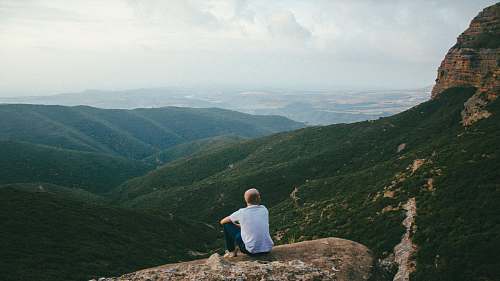 human person sitting on the edge of a cliff over looking mountains during daytime outdoors