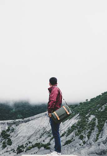 human man carrying black crossbody bag while standing on platform overlooking hill under white sky at daytime person