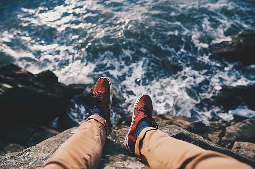human man in red sneakers sitting in cliff near body of water shoe