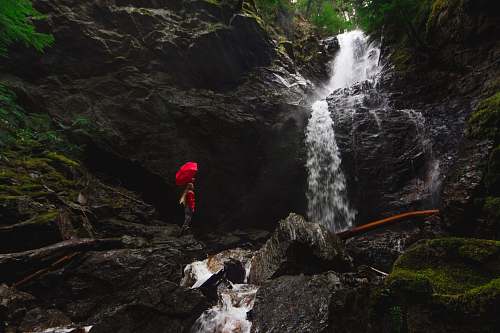 moss person standing on rock formation under red umbrella beside waterfalls waterfall