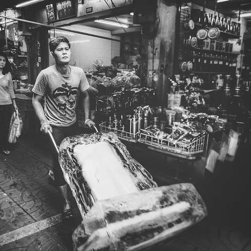 people grayscale photo of man carrying ice block using hand truck human