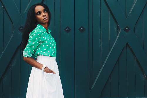 people woman in white skirt and green dress shirt hand on pocket standing at the front of green door person