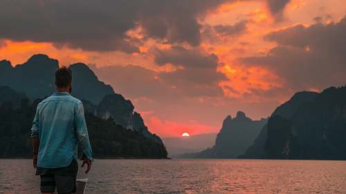person man standing near edge facing body of water during sunset human