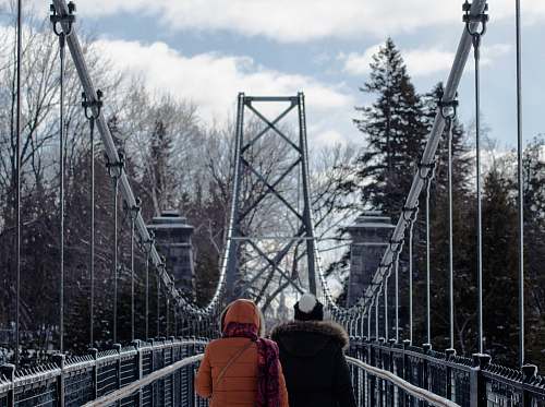 person two persons walking on gray suspension bridge in between trees at daytime human