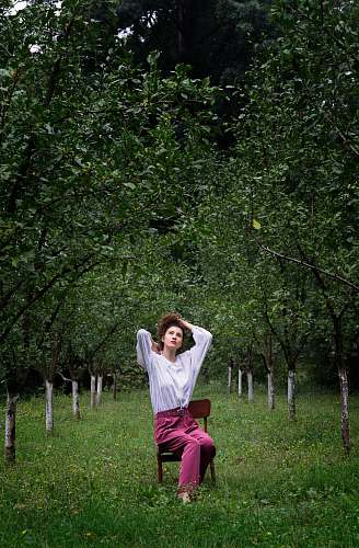 human woman sitting on brown armless chair while tying her hair under green trees at daytime person