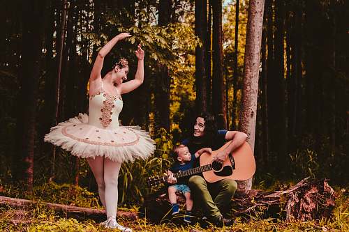 ballerina man sitting beside toddler boy while playing guitar and looking at the woman in ballerina dress in the woods performer