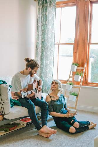 person man sitting on sofa playing guitar looking at girl sitting on the ground near window inside room people