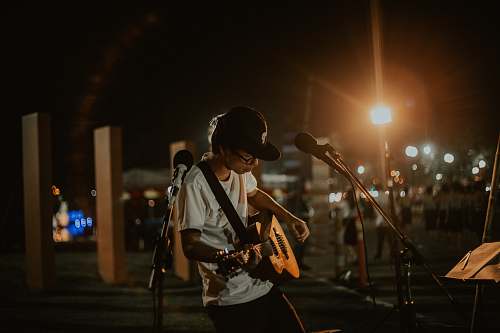 people man wearing white t-shirt playing guitar while standing in front of microphone with stand on open area during nighttime person