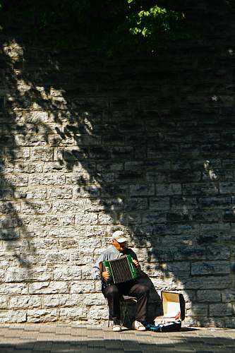 accordion man wearing grey shirt playing accordion instrument while sitting near concrete wall and case during daytime leisure activities