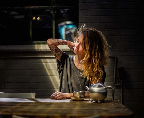 human woman in gray top sitting beside gray tea pot and cup on brown wooden table tattoo