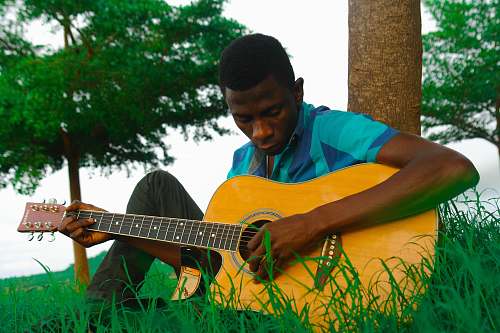 human man playing natural finish single-cutaway acoustic guitar while sitting on green grass near brown tree trunk during daytime people