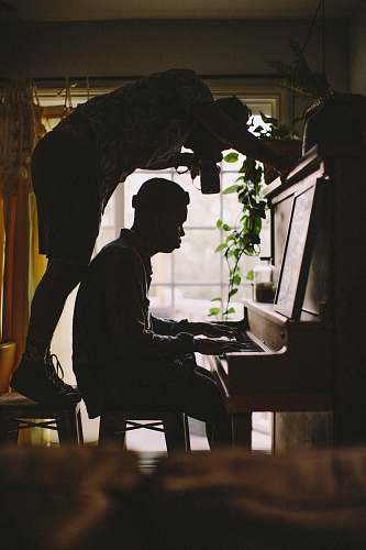 flora person sitting while playing piano while other man standing on stool while taking picture of person's hand on piano inside room jar