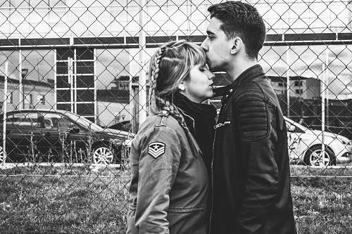 human grayscale photo of man kissing forehead of woman near fence black-and-white