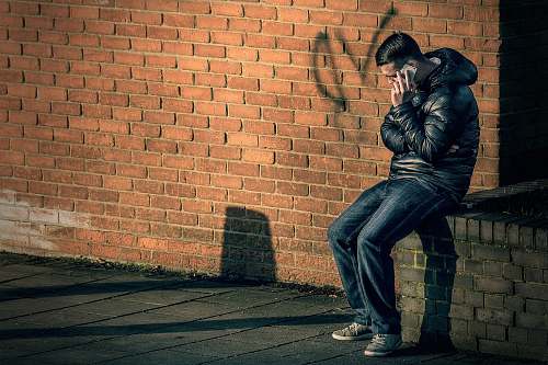 person man in black leather jacket sitting on brick wall holding smartphone human