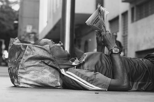 person man lying on sidewalk reading newspaper black-and-white