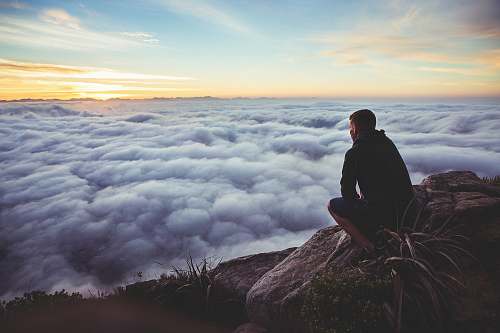 human man sitting on gray rock while staring at white clouds nature