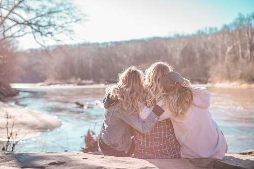 person three women sitting on rock near body of water surrounded by trees during daytime human