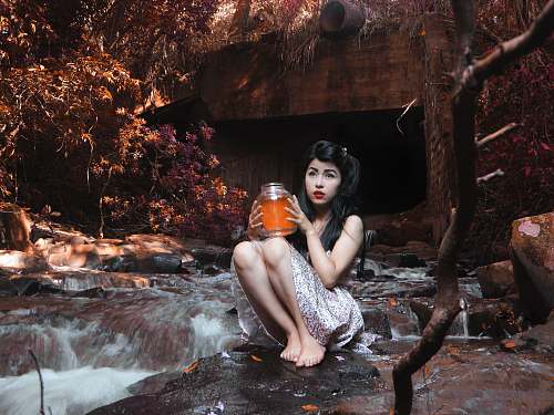 girl woman carrying glass jar while sitting on boulder beside flowing river behind brown bridge under shade of tree at daytime woman