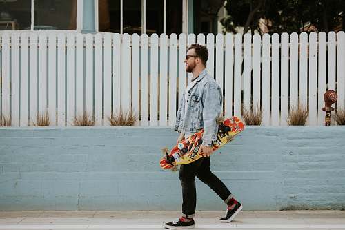 people man carrying long board walking beside white fence skateboard