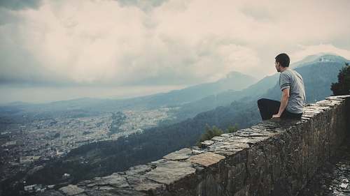 people man sitting alone on concrete brick wall facing mountain and city under cloudy sky human