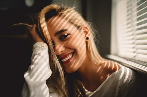 people selective focus photography of smiling woman holding her hair beside window blinds woman