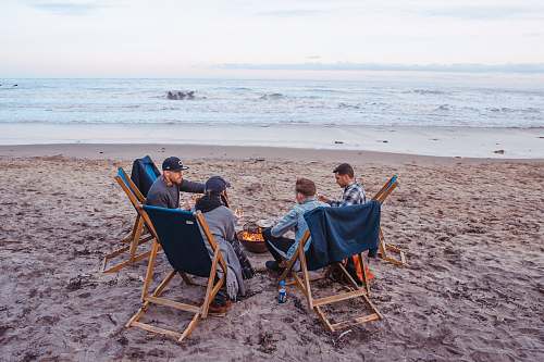 people three men and one woman sitting on beach lounge in front fire pit near seashore human