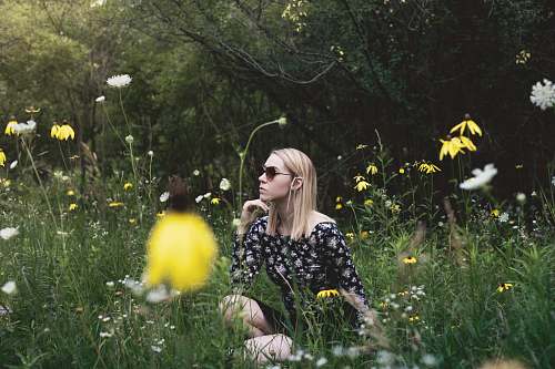 blonde woman sitting on grass field surrounded with flowers while hand on chin woman
