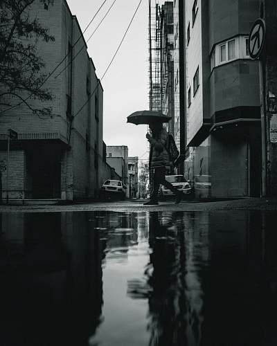 building greyscale photo of person walking and holding umbrella town