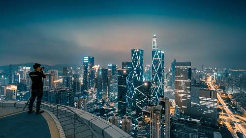 human man standing on high building taking photo of lighted high-rise buildings person