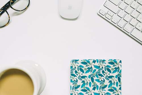 white Apple Magic keyboard with mouse; eyeglasses; notebook; white cup in flat lay photography desktop